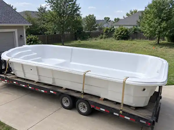 A white fiberglass pool shell sitting on a flatbed trailer in a driveway, with an unexcavated backyard visible behind it.