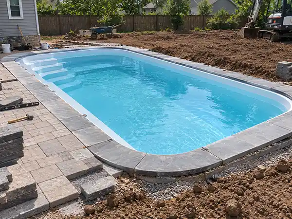 Pavers and coping being set around the edge of a finished fiberglass pool shell, with the yard still partly torn up.